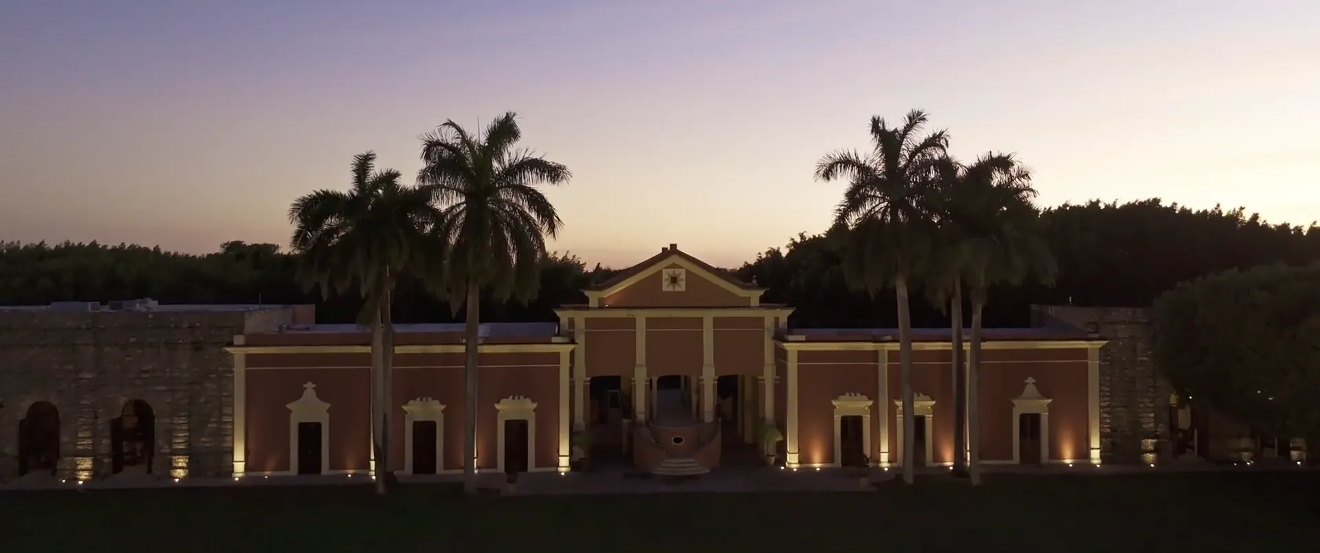 Hacienda Xtepen illuminated at dusk, framed by palm trees, in Mérida, Yucatán.