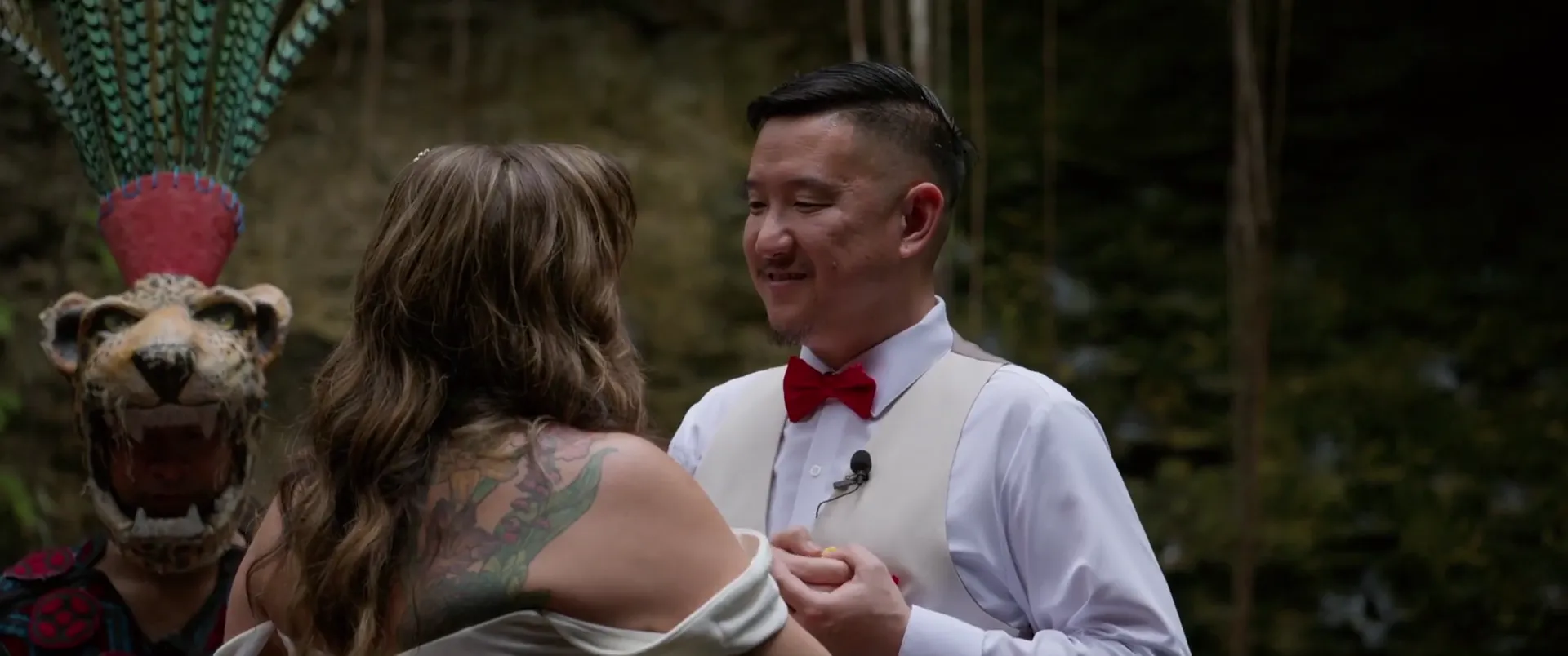 Maura y Jason tomados de las manos durante su ceremonia maya en el cenote, con un tocado ceremonial de jaguar visible detrás.