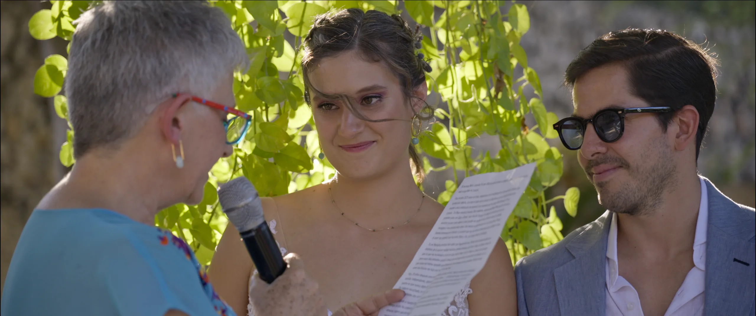 Wedding officiant holding a microphone as the bride reads her vows, with the groom listening.