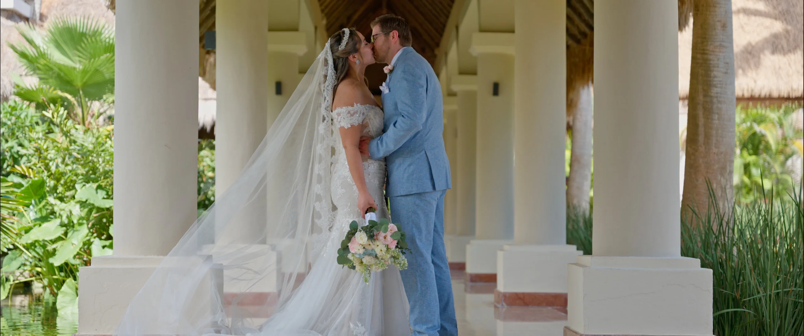Novia y novio besándose bajo una columnata junto a un estanque de lirios.