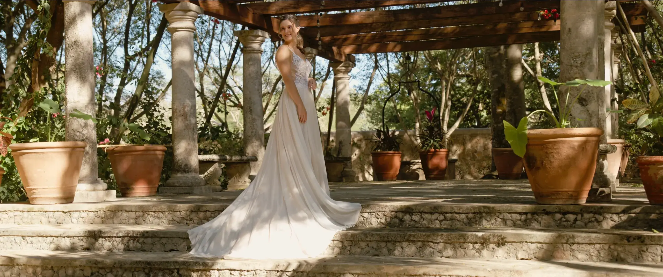 Novia con un vestido vaporoso de pie bajo una pérgola de piedra en un patio ajardinado.