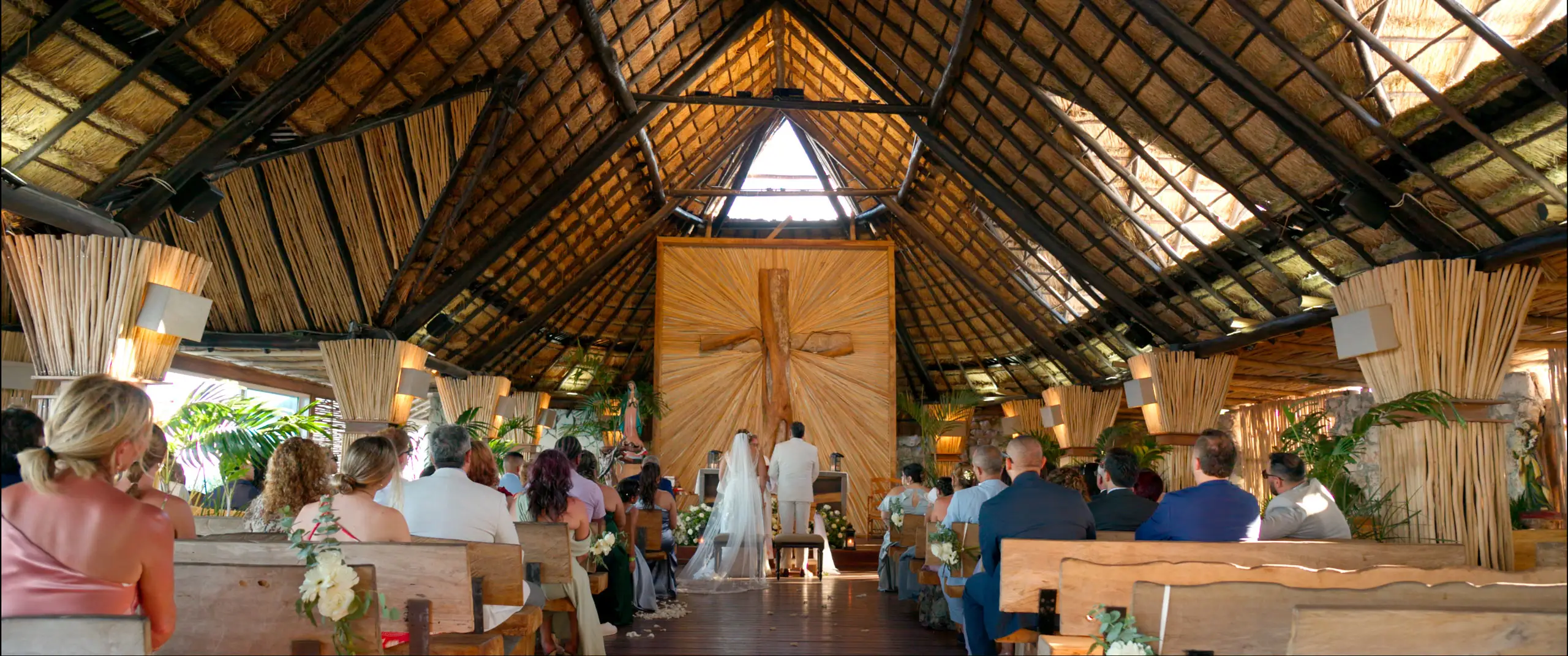 Ceremonia de boda dentro de una capilla con techo de palapa y una gran cruz de madera detrás del altar.