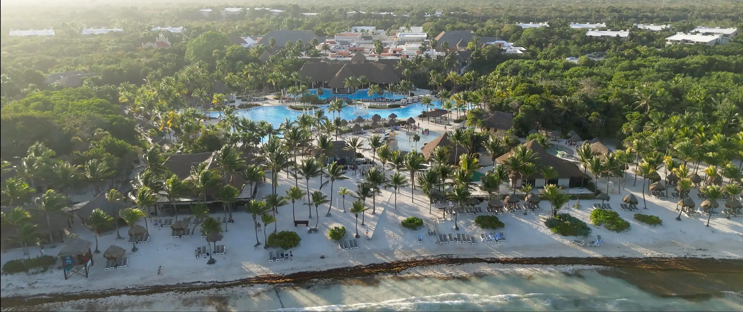 Aerial view of an all-inclusive beach resort with pools, palapas and palm trees along the Riviera Maya coastline.