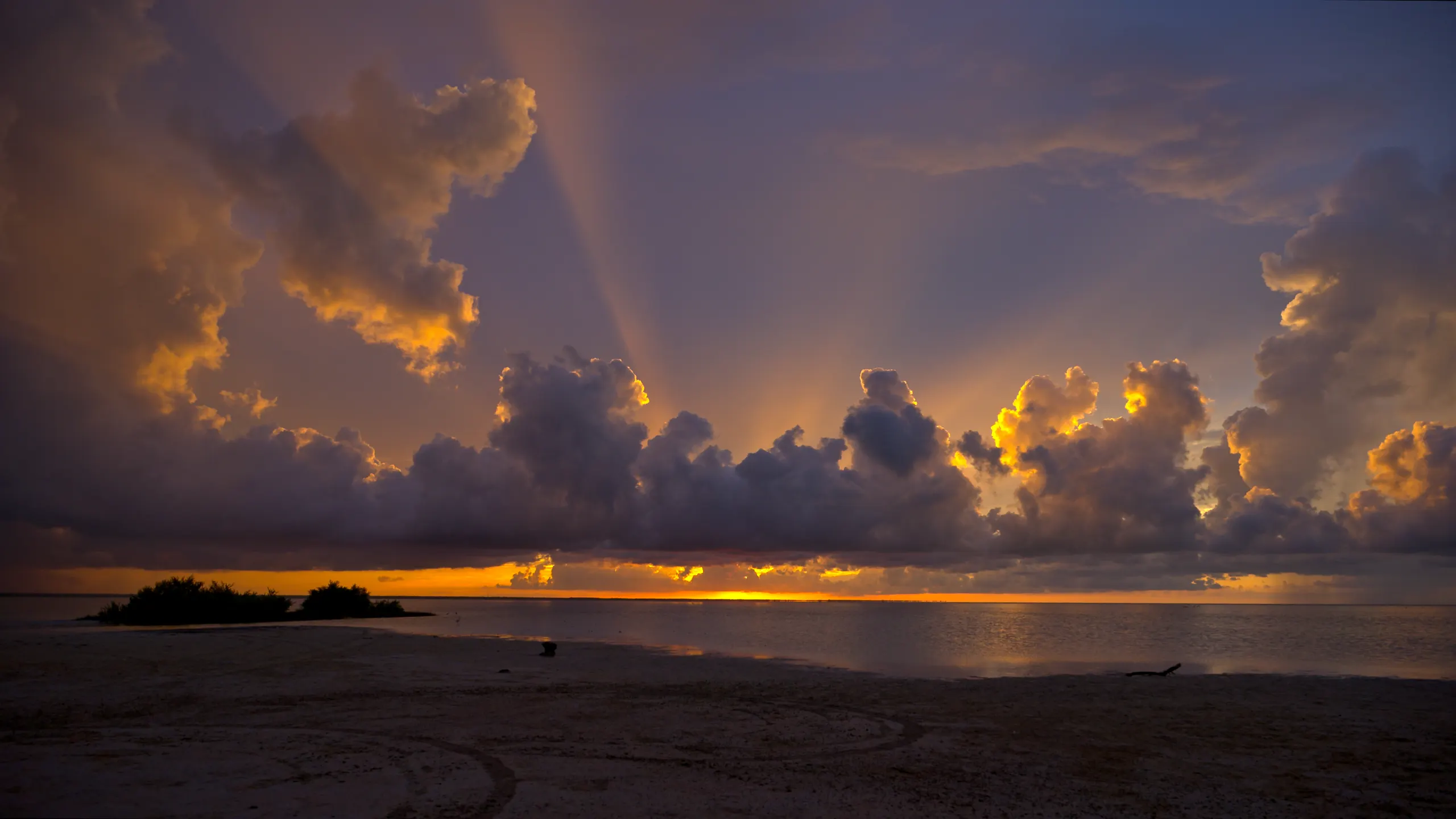 Atardecer dramático sobre la playa, con rayos de sol atravesando nubes de tormenta sobre el agua.