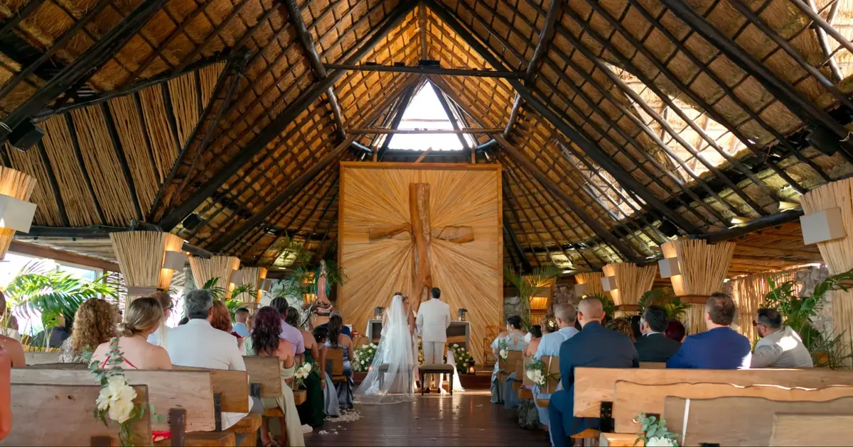 Ceremonia de boda dentro de una capilla con techo de palapa y una gran cruz de madera detrás del altar.