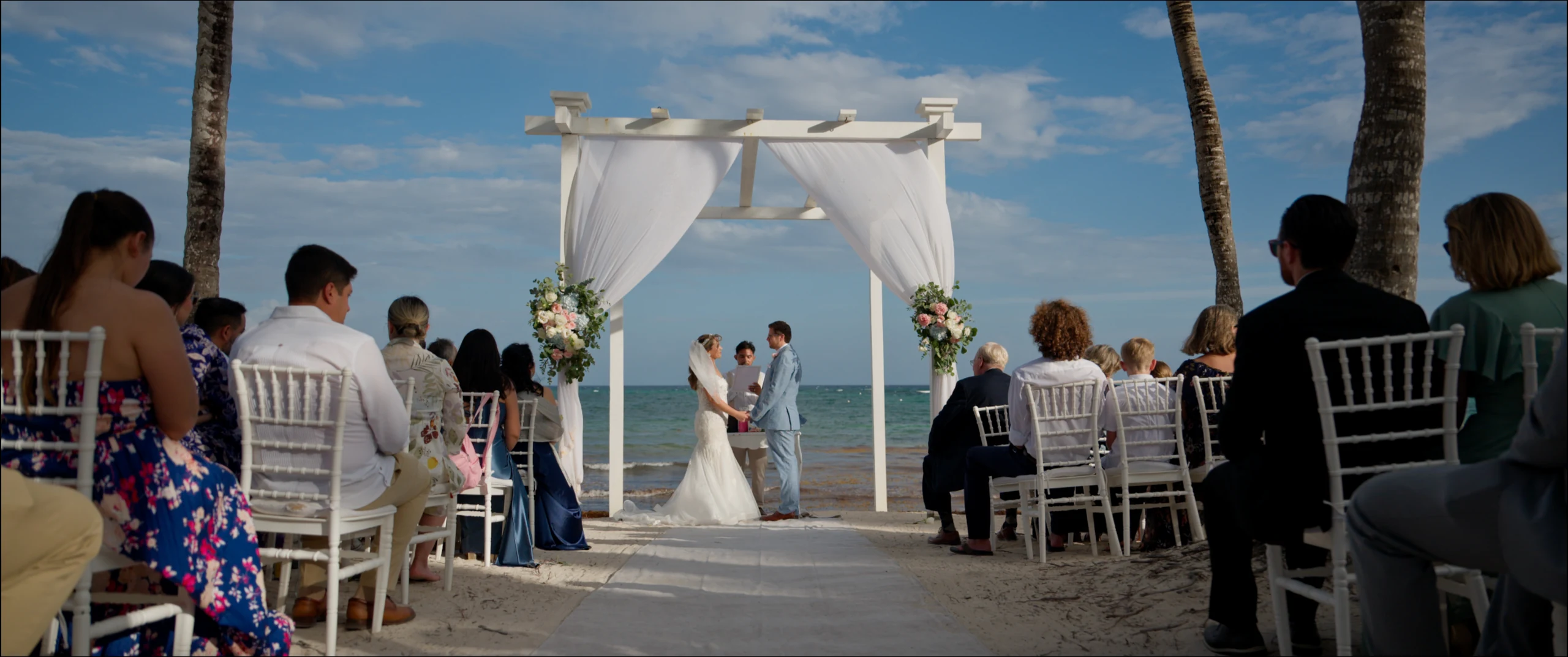 Diana and Zach exchanging vows at a beachfront altar at sunset at Grand Palladium Colonial, guests seated on the sand.