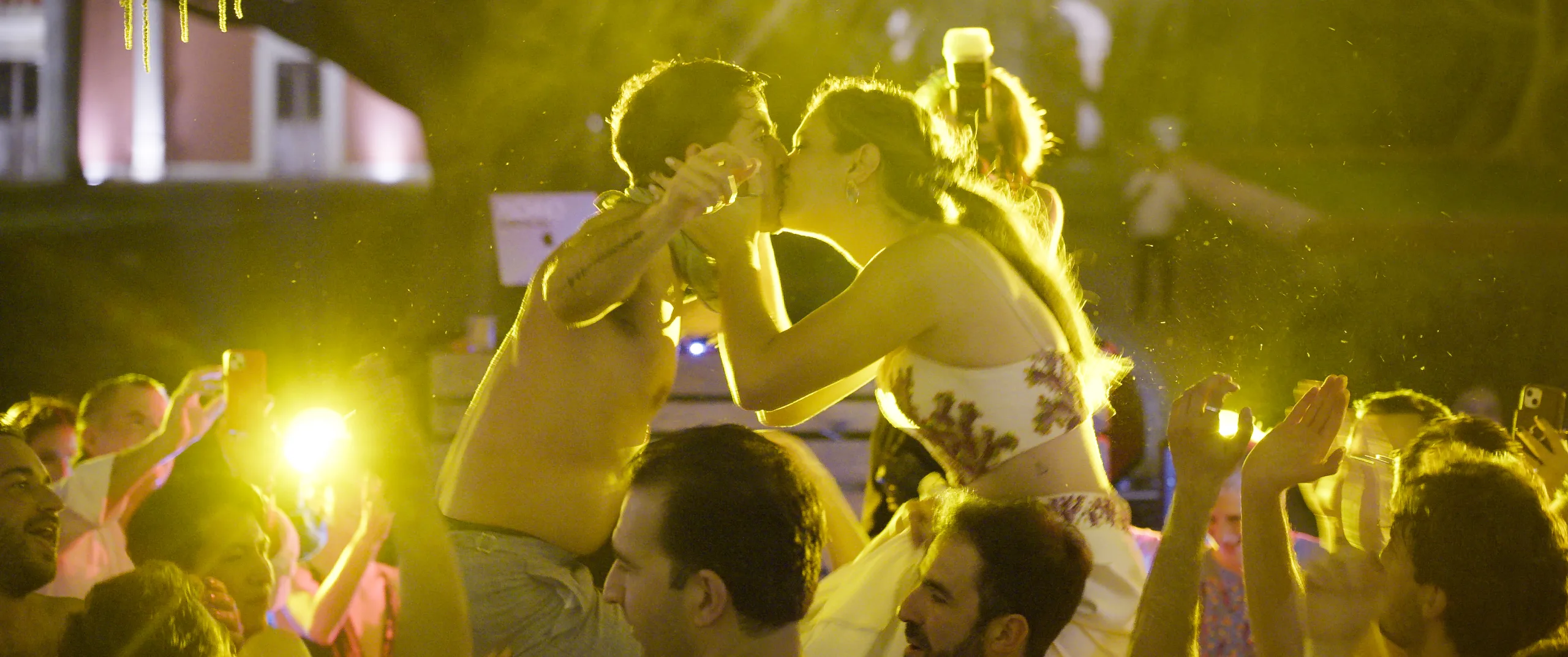 María and Fernando kissing while lifted on shoulders above the crowd during the batucada.