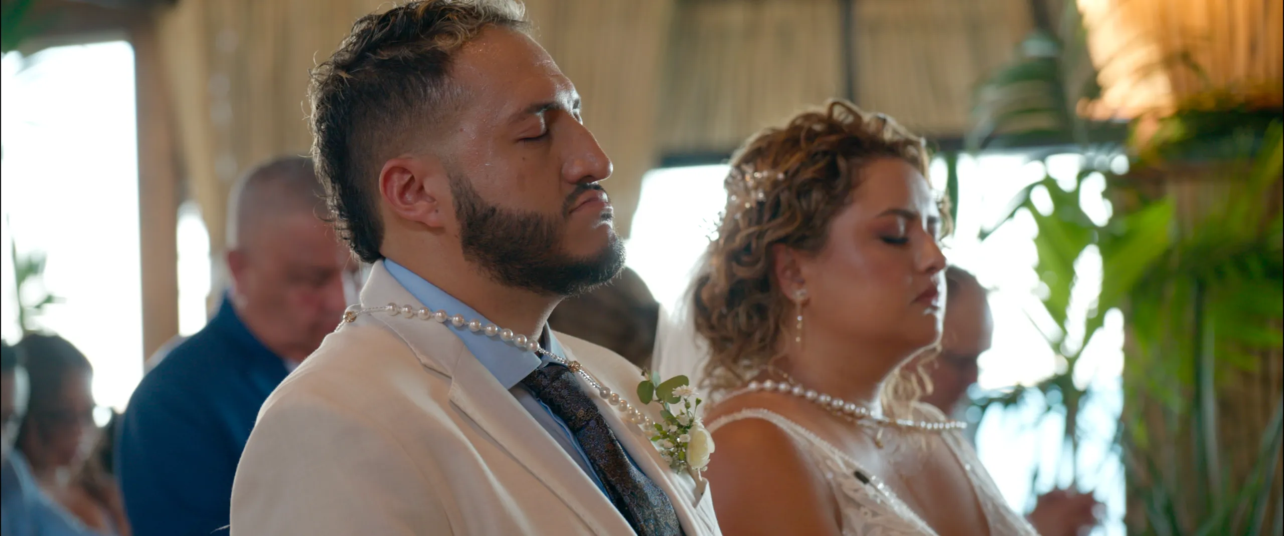 Novia y novio con los ojos cerrados durante una oración en su ceremonia católica, con los lazos de perlas tradicionales.