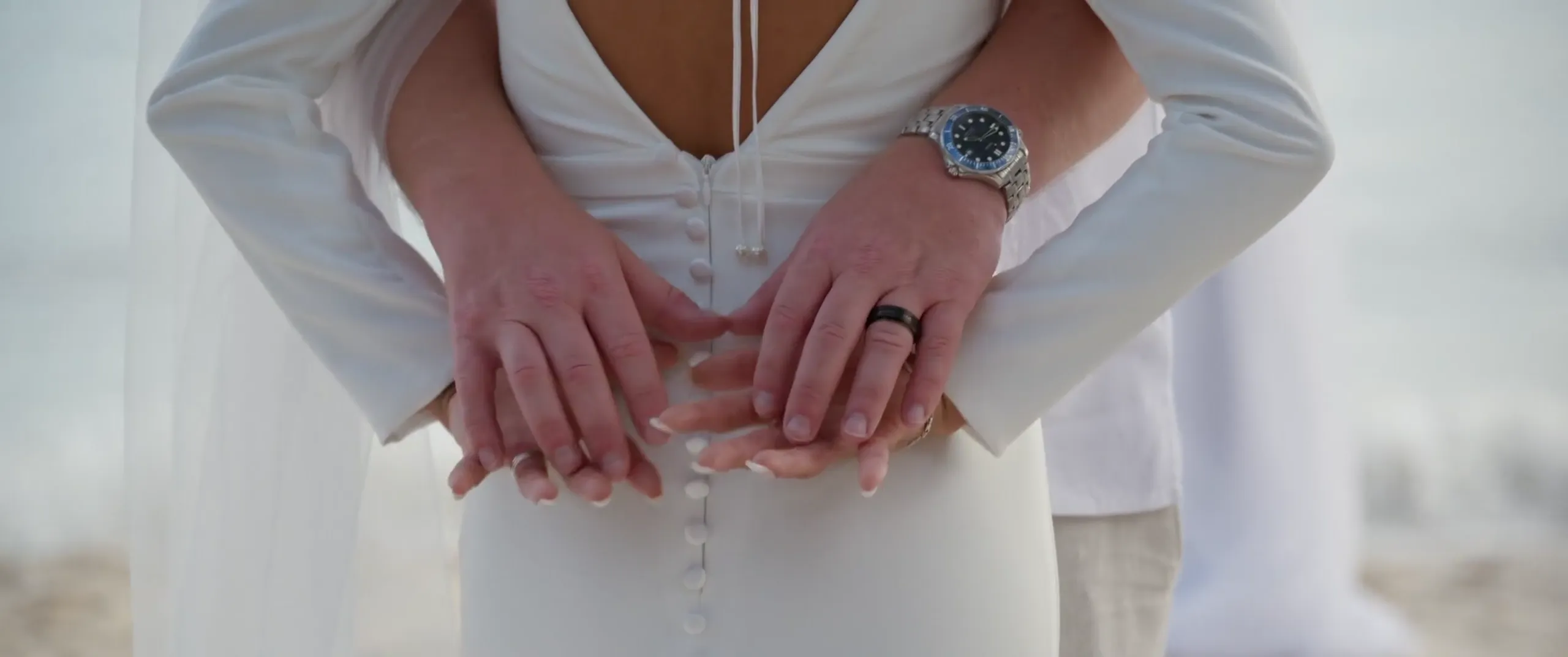 Primer plano del novio abrazando a la novia por la espalda, con las manos entrelazadas sobre el vestido de novia abotonado.