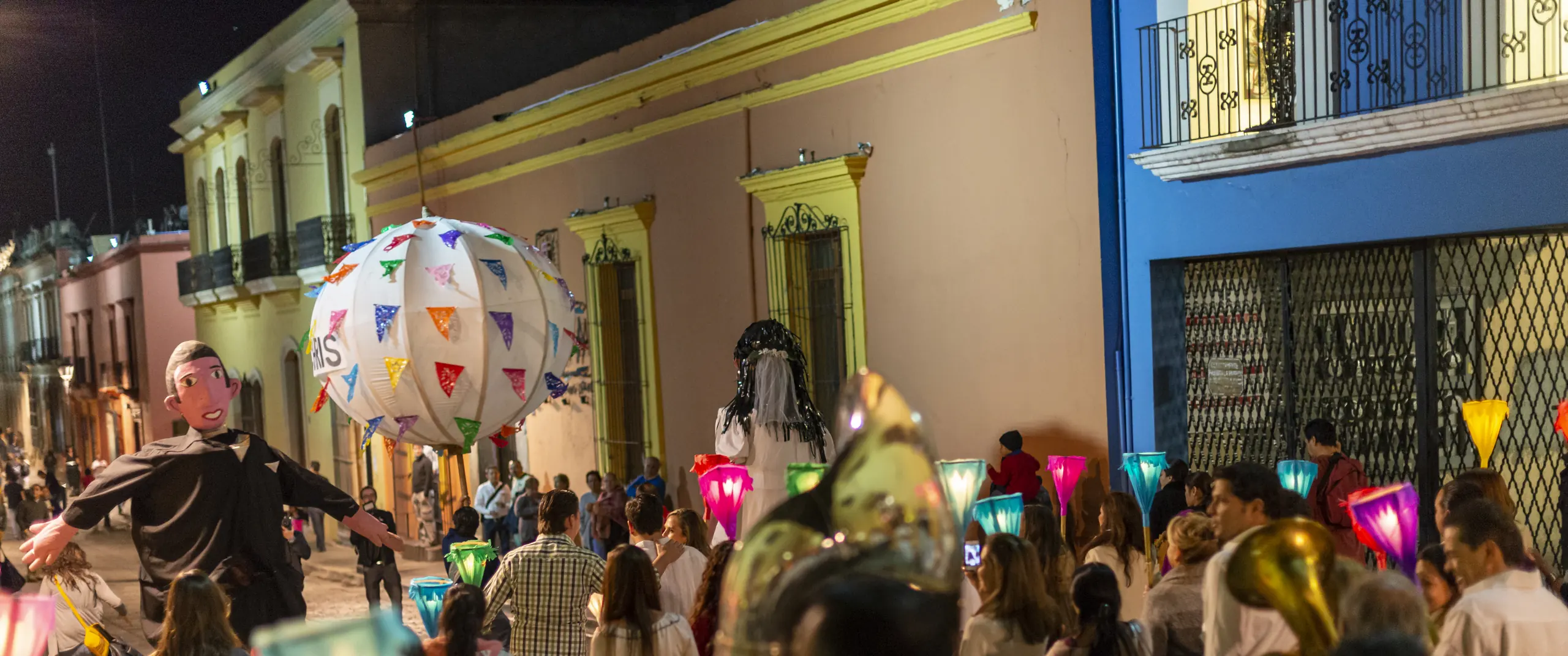 Una calenda de boda avanzando de noche por una calle colonial de Oaxaca, con mojigangas gigantes de los novios, una marmota iluminada, faroles de colores y una banda de viento.