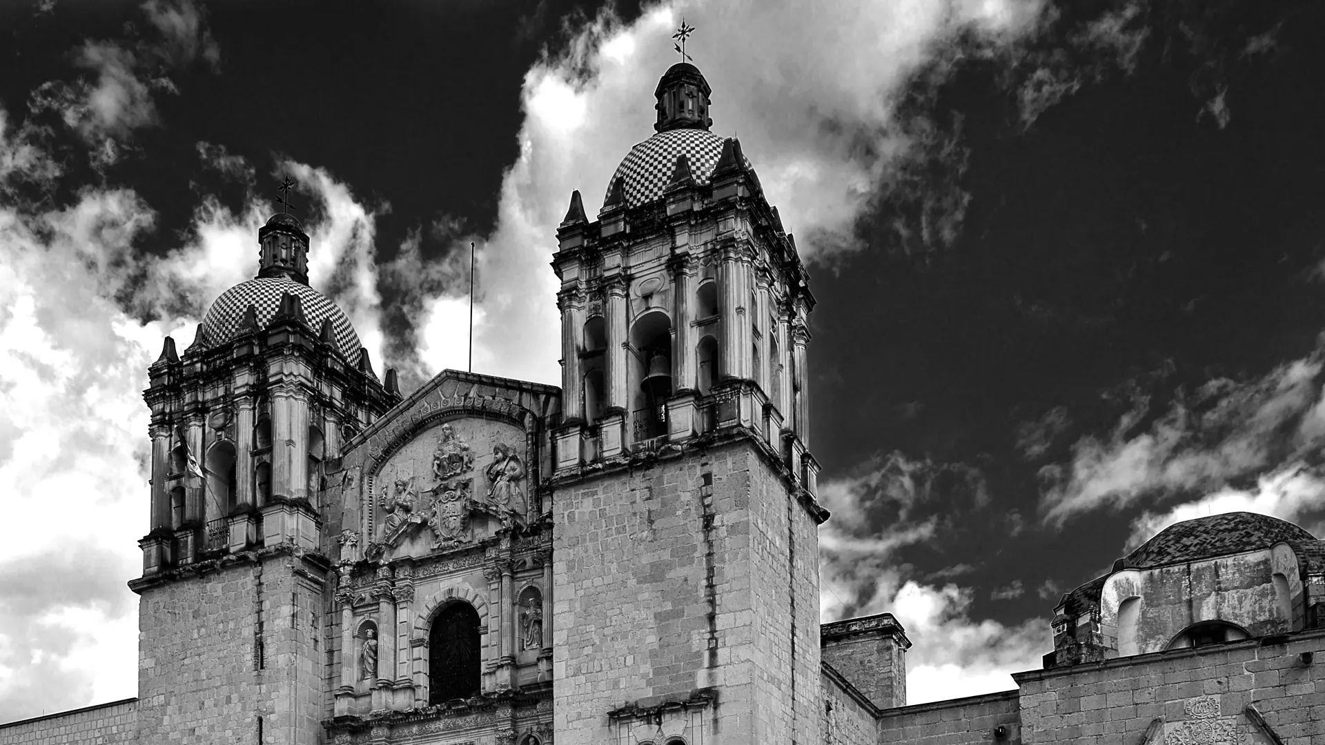 La fachada barroca y las torres gemelas de Santo Domingo de Guzmán, en Oaxaca, fotografiadas en blanco y negro contra un cielo dramático.